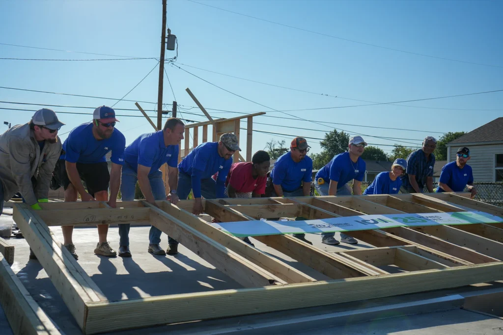 A group of volunteers working together to lift a large wooden wall frame on a concrete foundation.