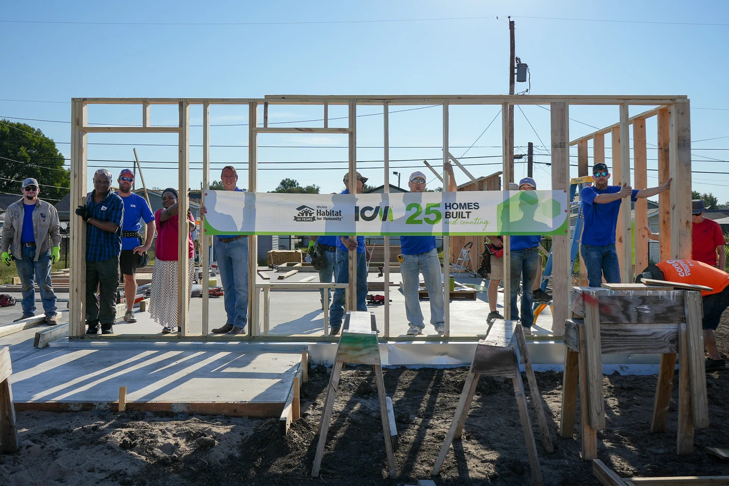 Volunteers standing behind a newly erected wooden wall frame, proudly displaying a Wichita Habitat for Humanity and ICM banner celebrating 25 homes built.