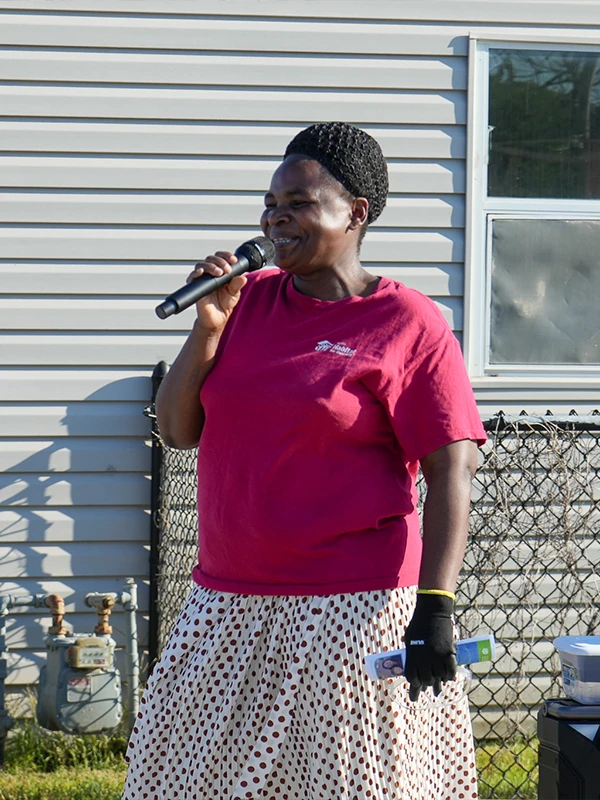 A woman in a red shirt and patterned skirt smiling while speaking into a microphone outdoors.