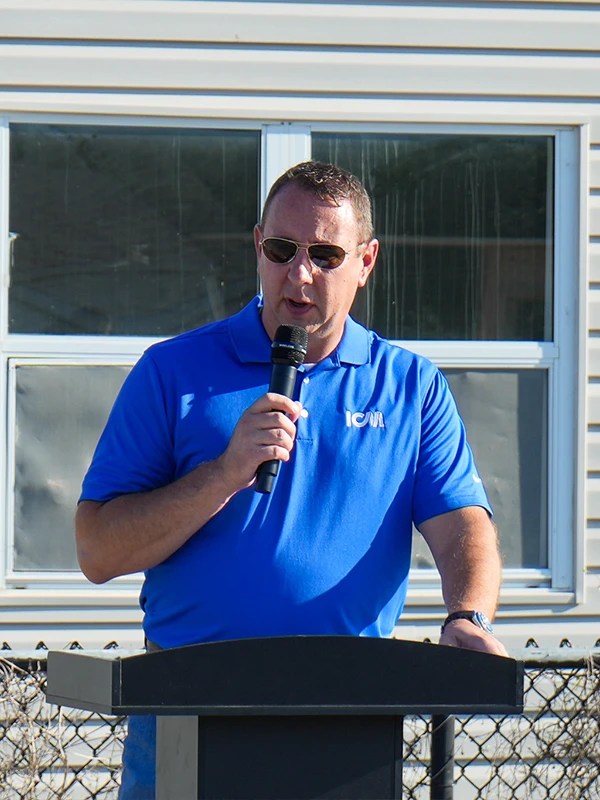 A man wearing a blue polo shirt and sunglasses speaking at a podium with a microphone.
