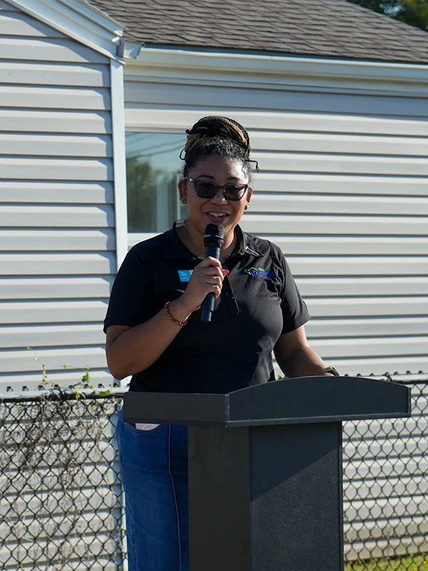 A woman wearing sunglasses and a black collared shirt smiling as she speaks at an outdoor podium.