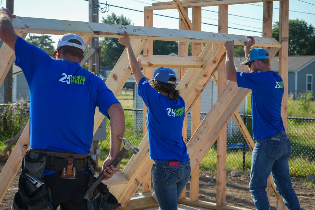 Three construction volunteers wearing matching blue shirts with "25 HOMES BUILT" printed on the back while lifting and steadying a wooden structure.