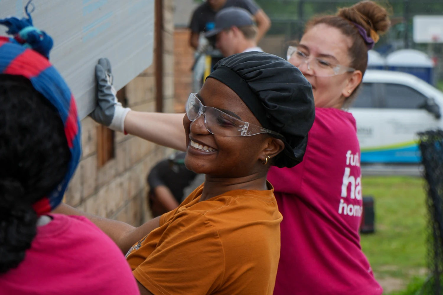 A smiling woman wearing safety glasses and a black hair wrap helps lift a large piece of construction material.