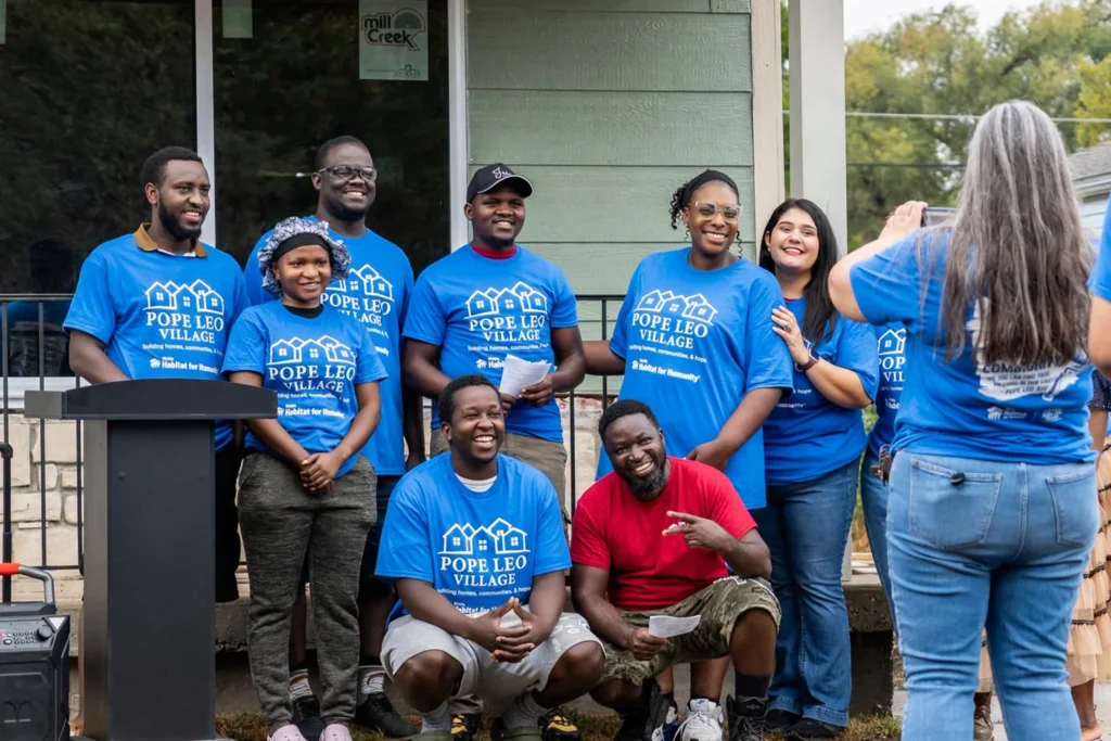 A group of eight people wearing matching blue Pope Leo Village t-shirts smile while posing for a photo in front of a house.