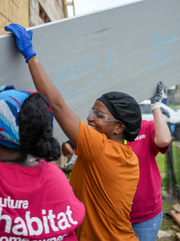 Three volunteers lift a large grey construction board together at a building site.
