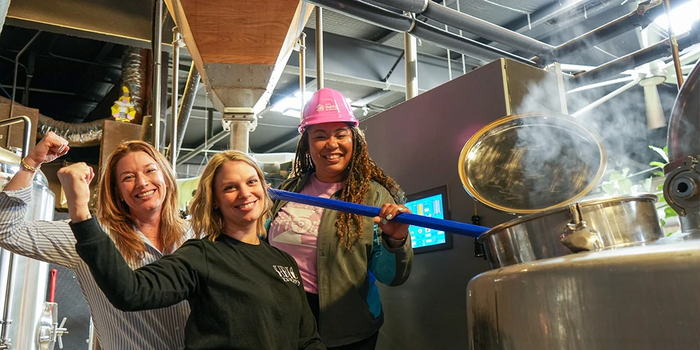 Three women posing playfully while brewing beer, including one in a pink Habitat hard hat.