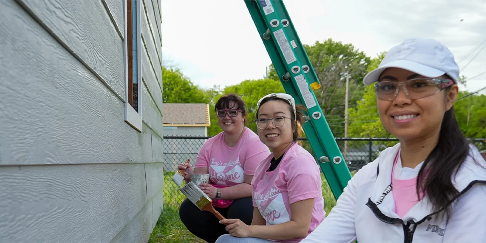 Three smiling volunteers in pink and white attire painting the siding of a house outdoors.