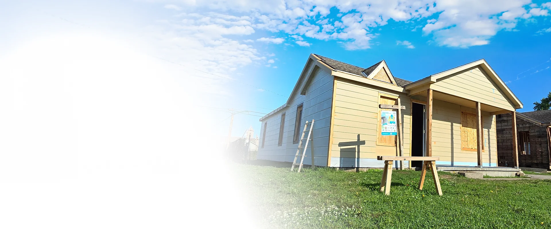 A single-story house under construction with light yellow siding on a grassy lawn under a blue sky, with a wooden sawhorse in the foreground.