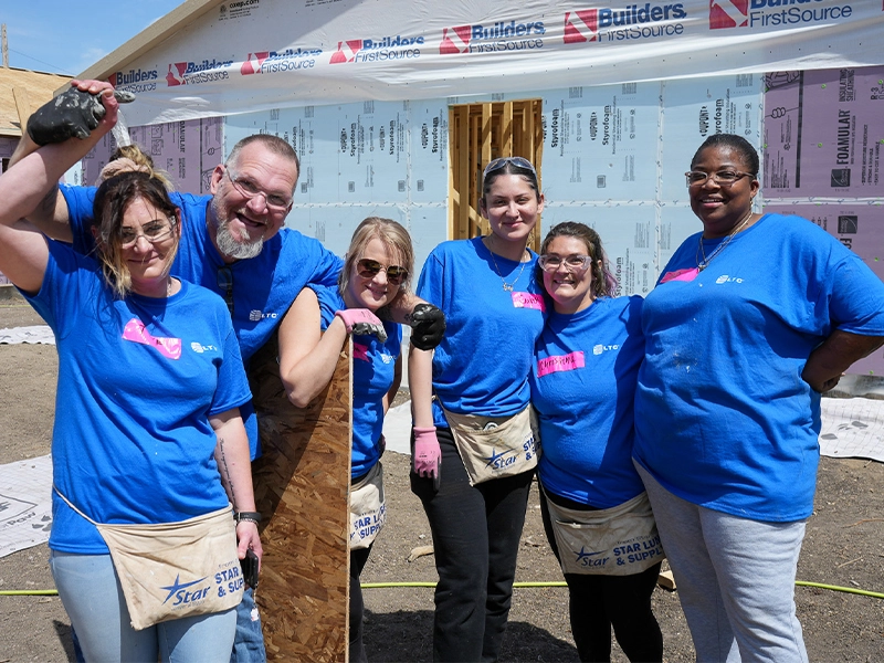 Six volunteers posing for a group photo in front of a house under construction, wearing blue shirts and tool aprons.
