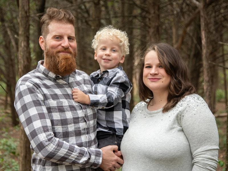 Family partners Seth, Krista, and their son taking a family photo in front of trees.