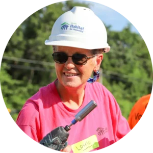 Close-up of a smiling volunteer named Lorrie wearing a hard hat and holding a drill.